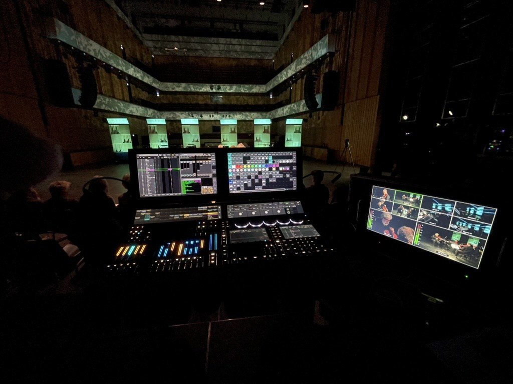 Lysbord med to skjermer og en videomonitor foran en konsertsal med opplyste paneler, under live-innspilling med orkester